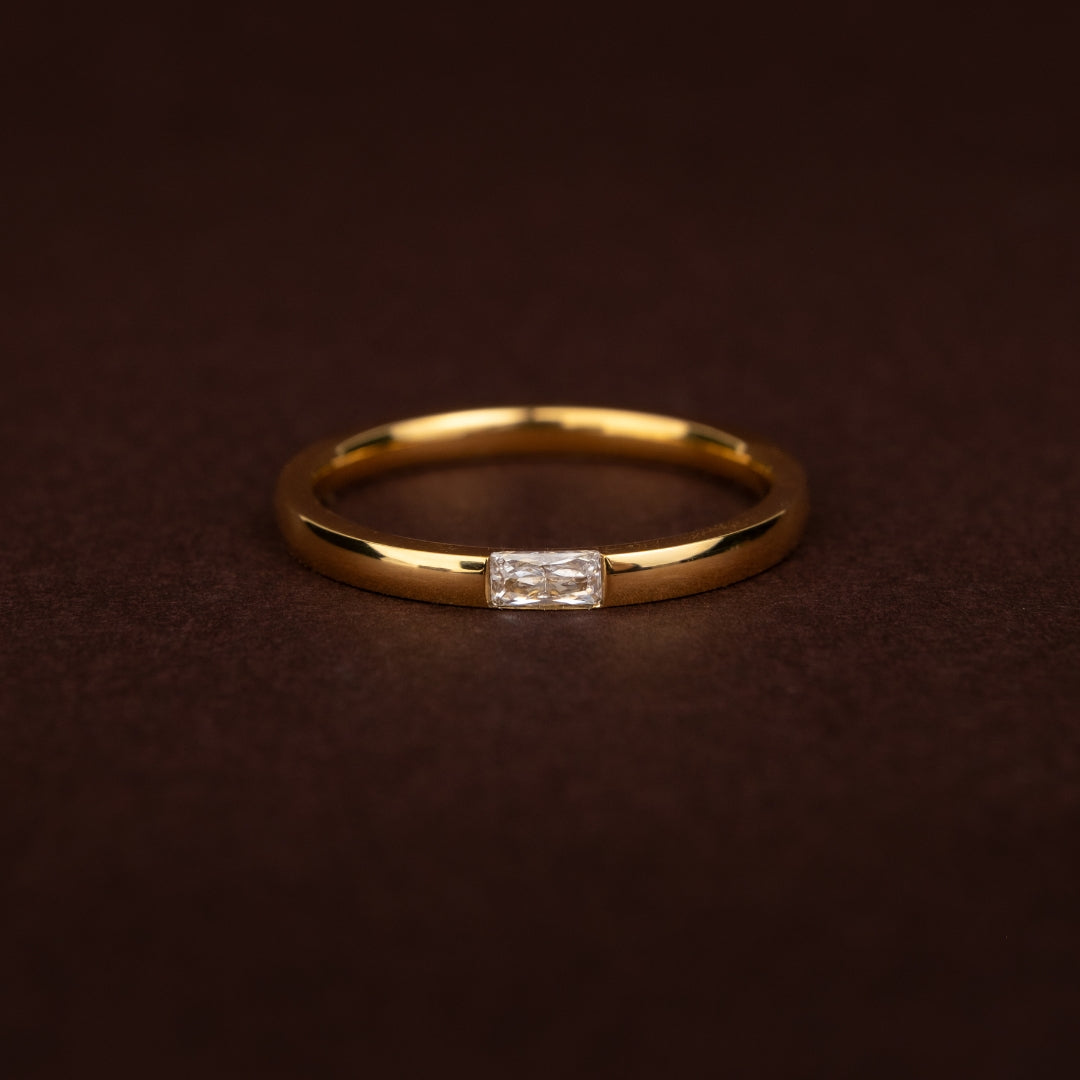 Anel dourado fino e minimalista em aço inoxidável com uma pequena pedra retangular (zircónia baguette) ao centro sobre fundo escuro.