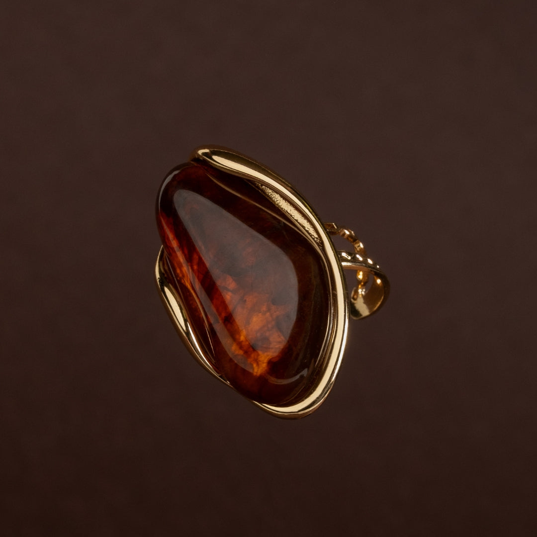 Anel dourado grande com pedra orgânica escura tom âmbar ou cognac e aro fluido em aço inoxidável sobre fundo escuro.