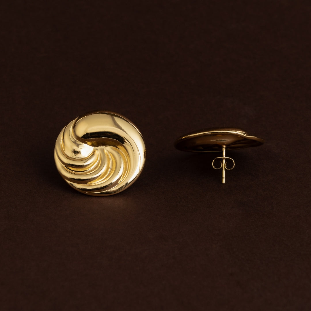 Brincos de botão dourados grandes em aço inoxidável com textura em relevo espiral ou turbina, estilo vintage sobre fundo escuro.