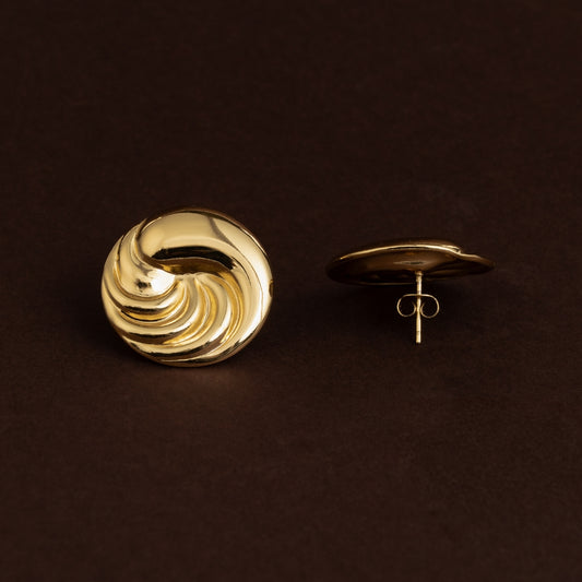 Brincos de botão dourados grandes em aço inoxidável com textura em relevo espiral ou turbina, estilo vintage sobre fundo escuro.