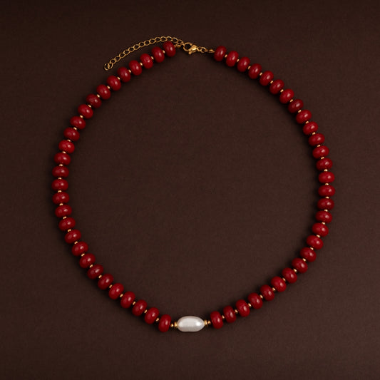 Colar curto de contas redondas vermelhas vivas com espaçadores dourados e uma pérola barroca branca no centro sobre fundo escuro.