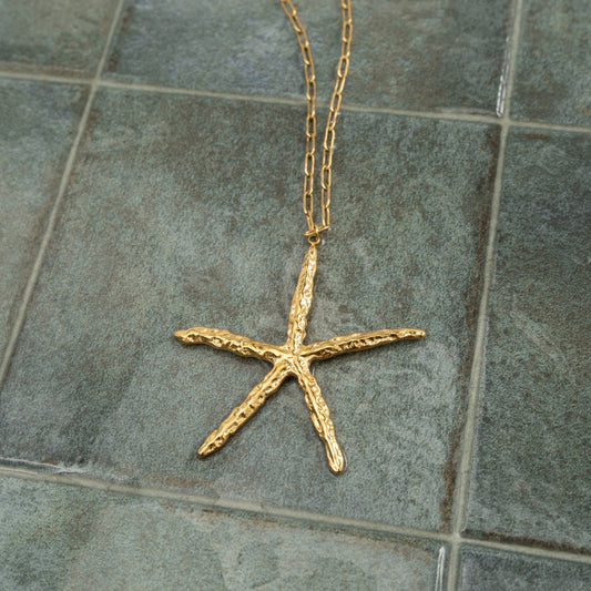 Colar Estrela do Mar longo em aço inoxidável dourado com textura irregular estendido sobre azulejo verde