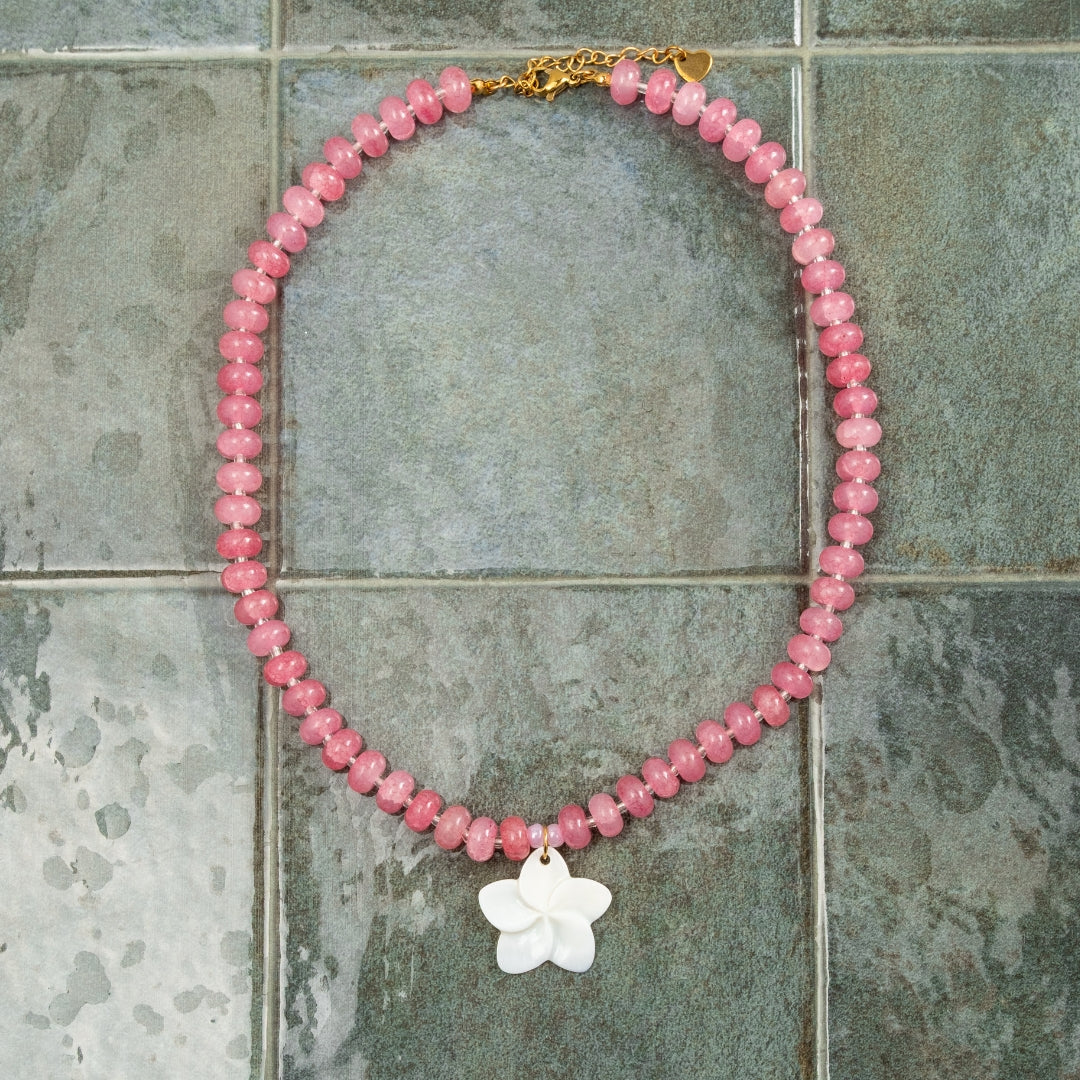Colar Aloha em aço inoxidável dourado com pedras naturais rosa e flor branca, estendido sobre fundo de azulejo verde