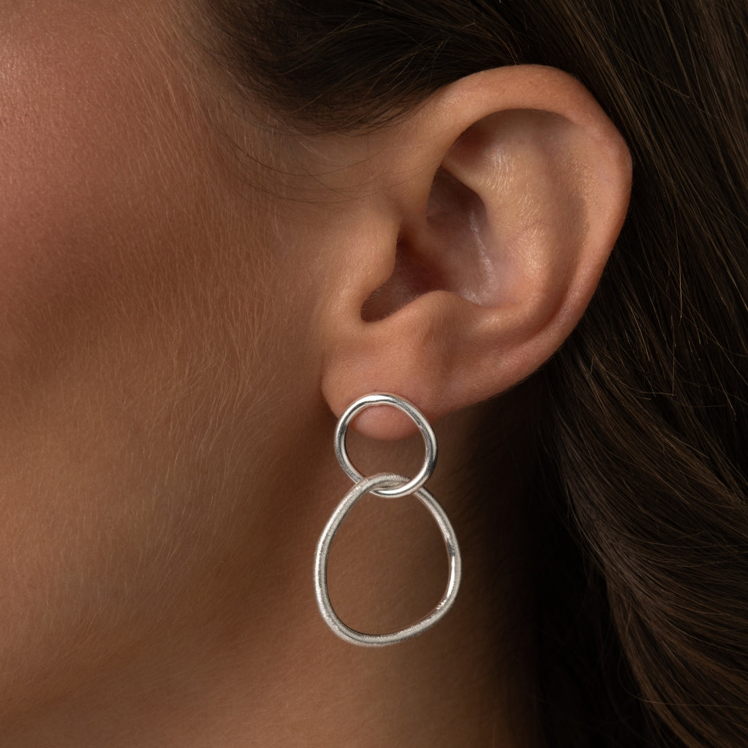 Close-up de Brincos Dualidade em Prata de Lei 925 com dois elos interligados, um circular e um de forma orgânica e abstrata, usados na orelha de uma mulher.