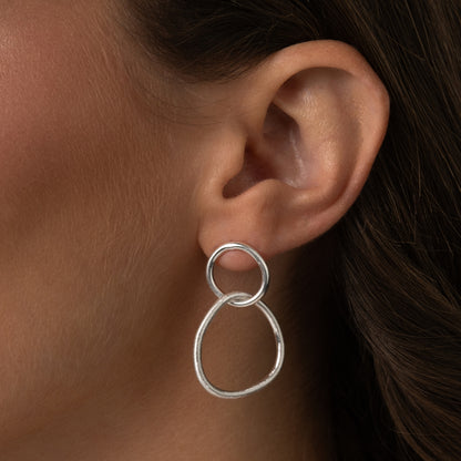 Close-up de Brincos Dualidade em Prata de Lei 925 com dois elos interligados, um circular e um de forma orgânica e abstrata, usados na orelha de uma mulher.