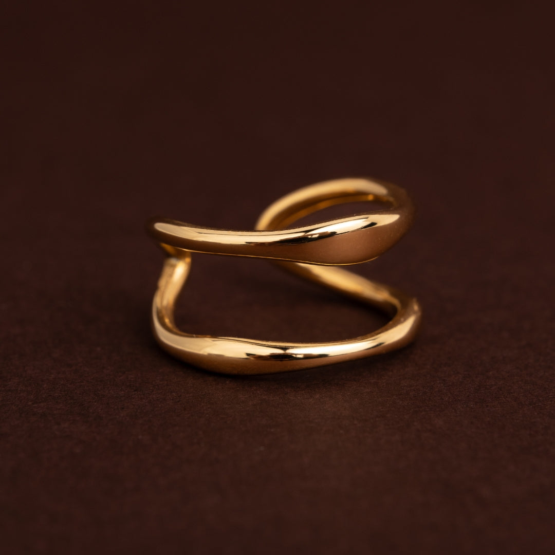 Anel dourado em aço inoxidável com duas bandas onduladas paralelas e espaço vazio no centro, estilo fluido e orgânico sobre fundo escuro.
