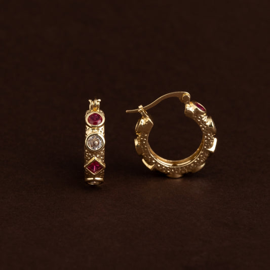 Par de argolas douradas com textura pontilhada e pedras zircónia transparentes e rubi cravadas, estilo vintage sobre fundo escuro.