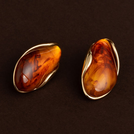 Par de brincos de botão dourados com pedra grande de resina efeito âmbar (castanho translúcido) e moldura orgânica em aço inoxidável sobre fundo escuro.
