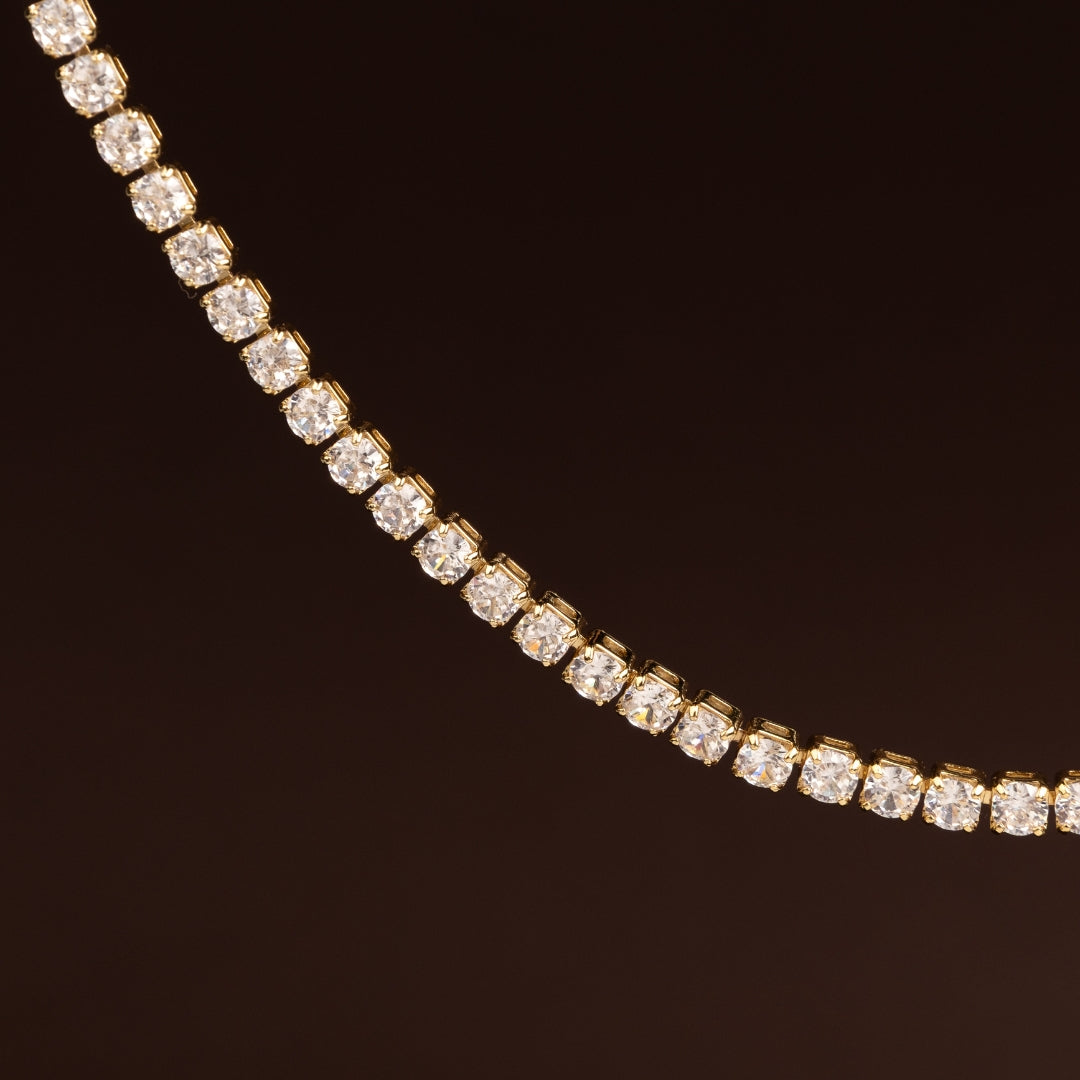Colar estilo tennis ou rivière com linha contínua de cristais quadrados/redondos brilhantes em base dourada sobre fundo escuro.
