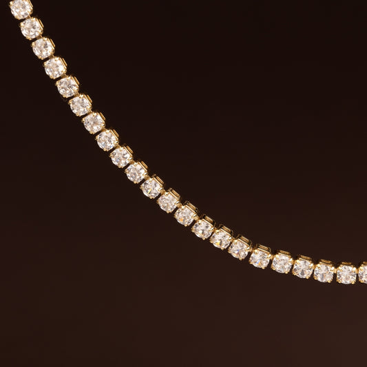 Colar estilo tennis ou rivière com linha contínua de cristais quadrados/redondos brilhantes em base dourada sobre fundo escuro.