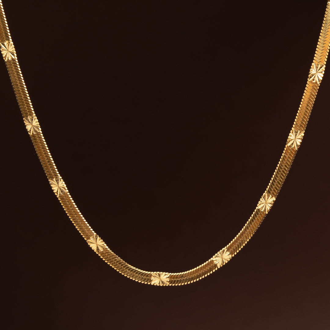 Imagem macro de Corrente dourada plana em aço inoxidável com padrão texturado de estrelas ou raios de sol gravados, estilo malha brilhante sobre fundo escuro.