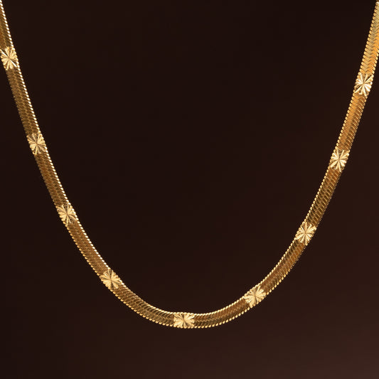 Imagem macro de Corrente dourada plana em aço inoxidável com padrão texturado de estrelas ou raios de sol gravados, estilo malha brilhante sobre fundo escuro.