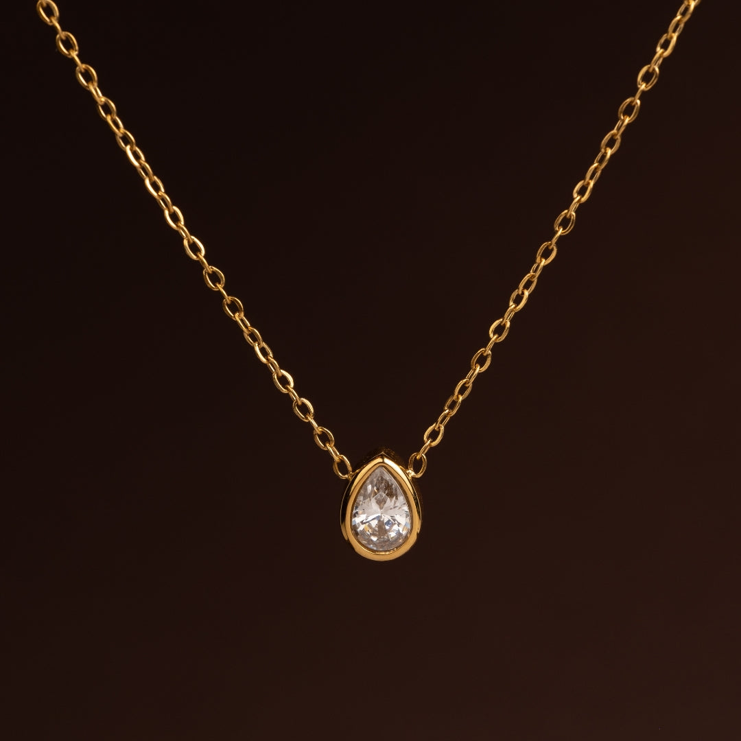 Colar dourado de corrente fina com um pendente solitário de cristal transparente em formato de gota ou pêra (teardrop) sobre fundo escuro.