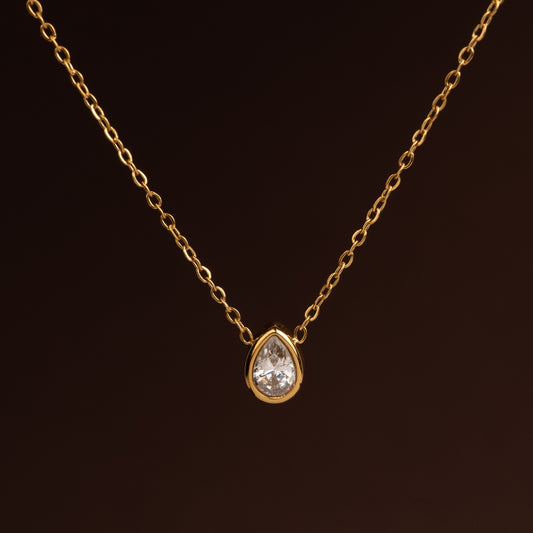 Colar dourado de corrente fina com um pendente solitário de cristal transparente em formato de gota ou pêra (teardrop) sobre fundo escuro.