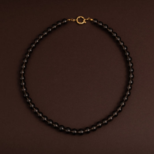 Colar de contas redondas translúcidas em tom castanho acinzentado escuro (quartzo fumado) com fecho boia dourado ao centro sobre fundo escuro.