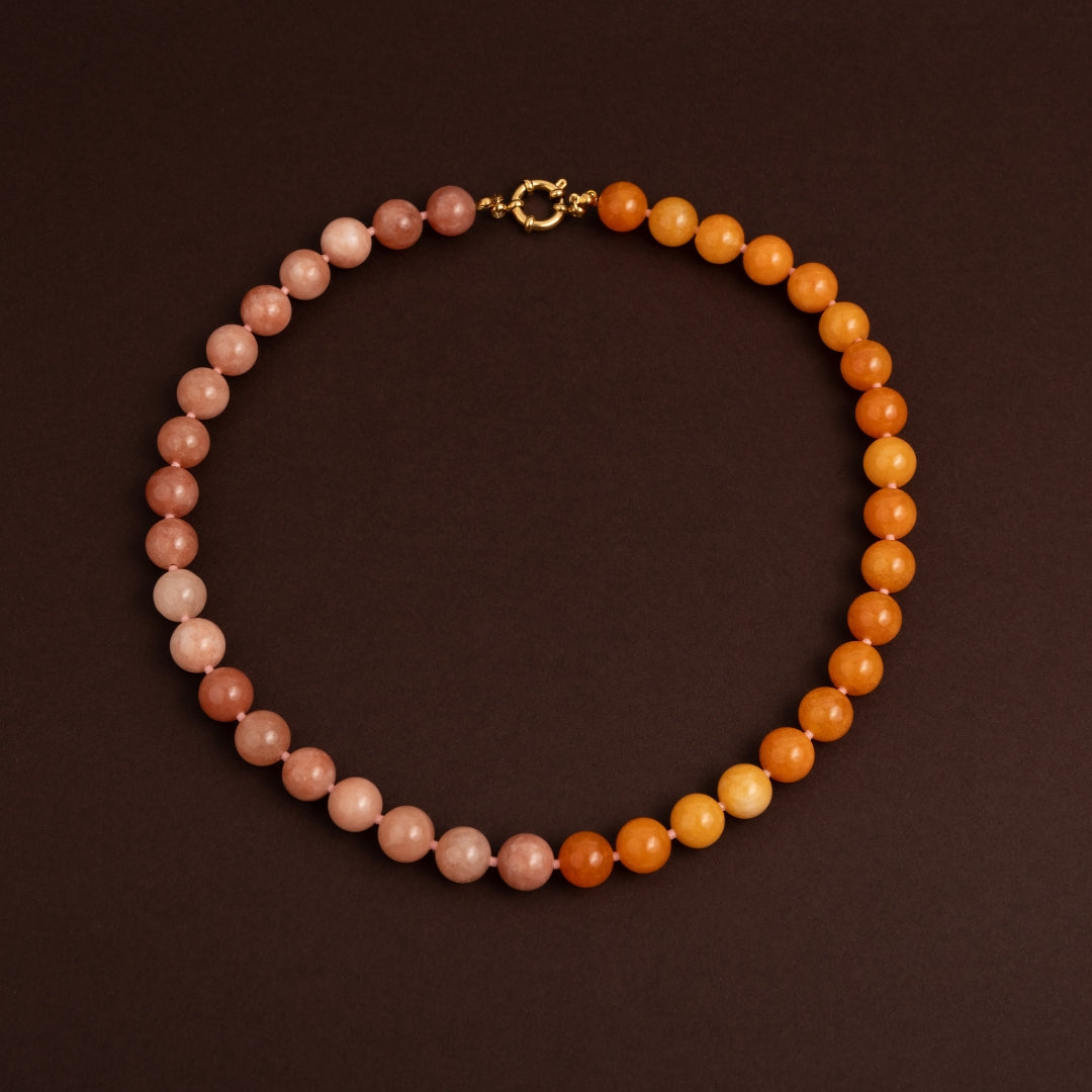Colar de contas redondas de pedra natural com gradiente de cor (dégradé) do rosa claro ao laranja escuro sobre fundo castanho.