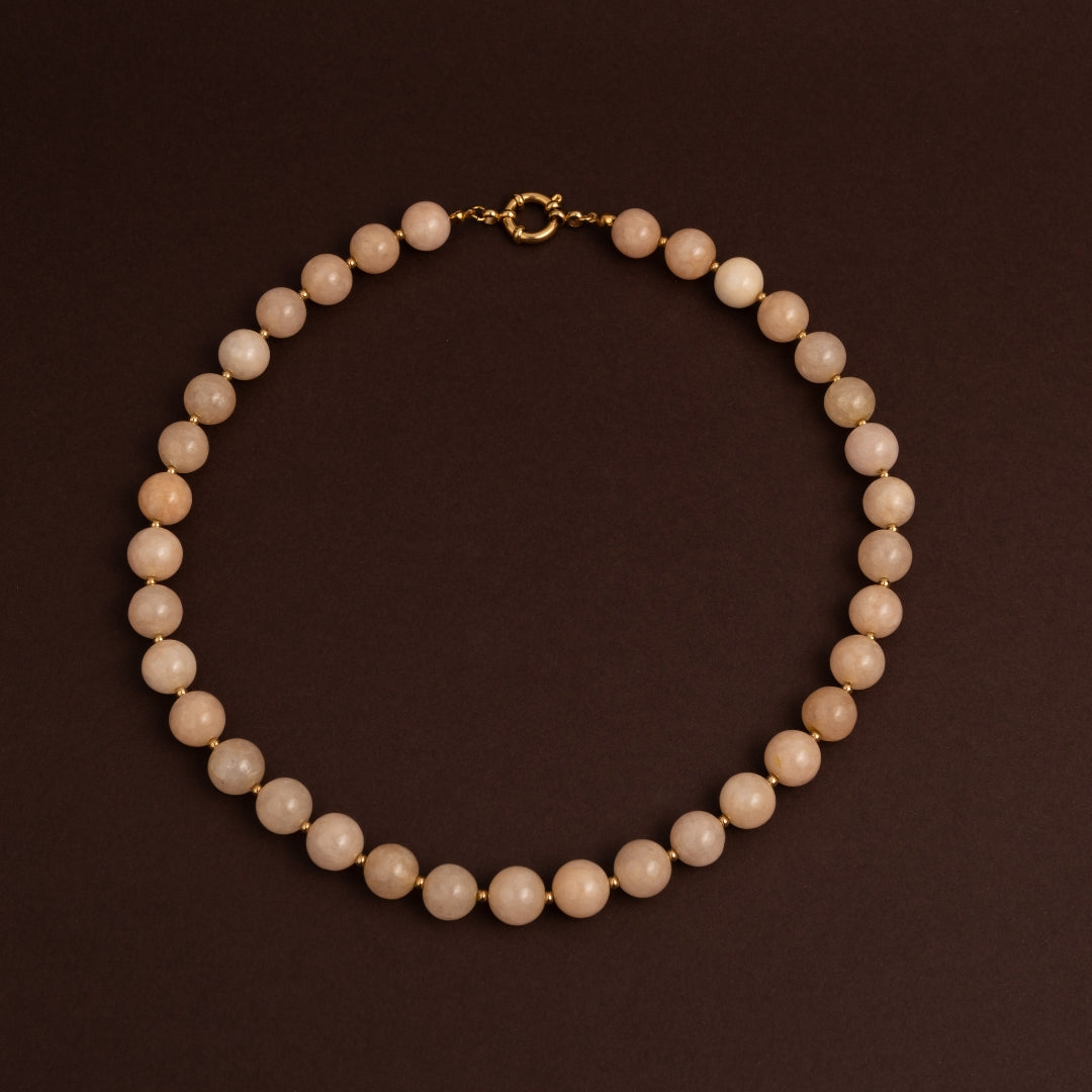 Colar de contas redondas de pedra natural em tom nude ou pêssego suave com fecho boia dourado ao centro sobre fundo escuro.