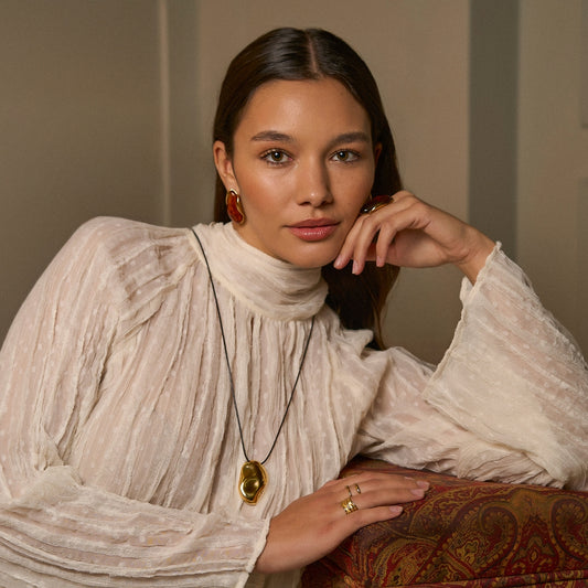 Modelo a usar colar escultural comprido com pendente dourado grande sobre blusa branca, estilo artístico e boémio.