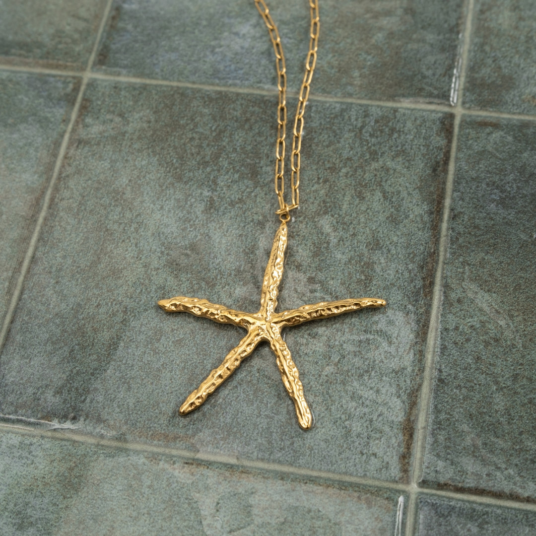 Colar Estrela do Mar longo em aço inoxidável dourado com textura irregular estendido sobre azulejo verde