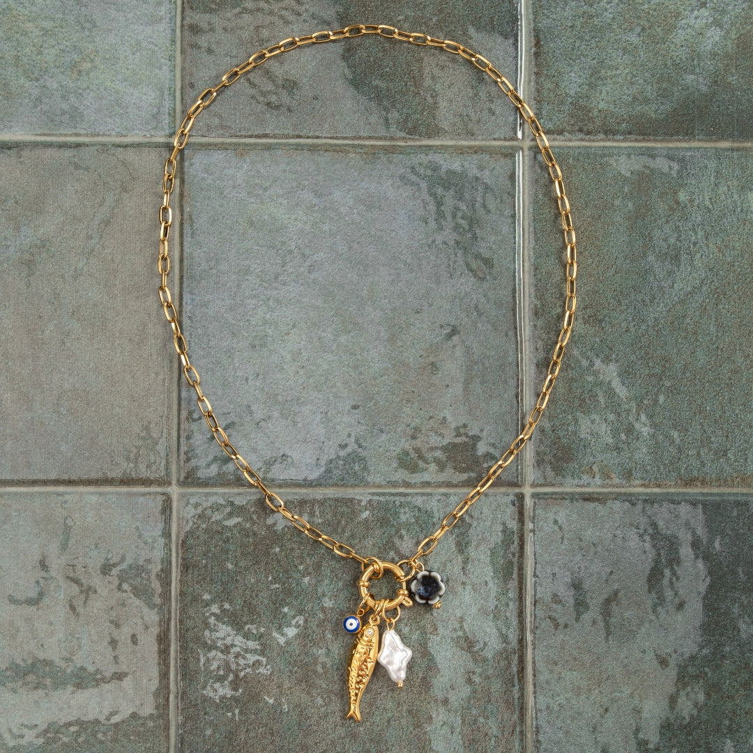 Colar Costa Nova dourado com amuletos de verão estendido sobre fundo de azulejo verde