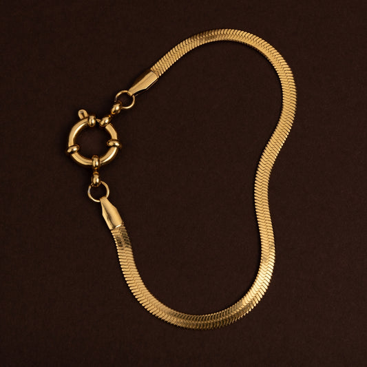 Pulseira dourada de malha plana e lisa (tipo fita ou herringbone) com fecho boia redondo grande em aço inoxidável sobre fundo escuro.