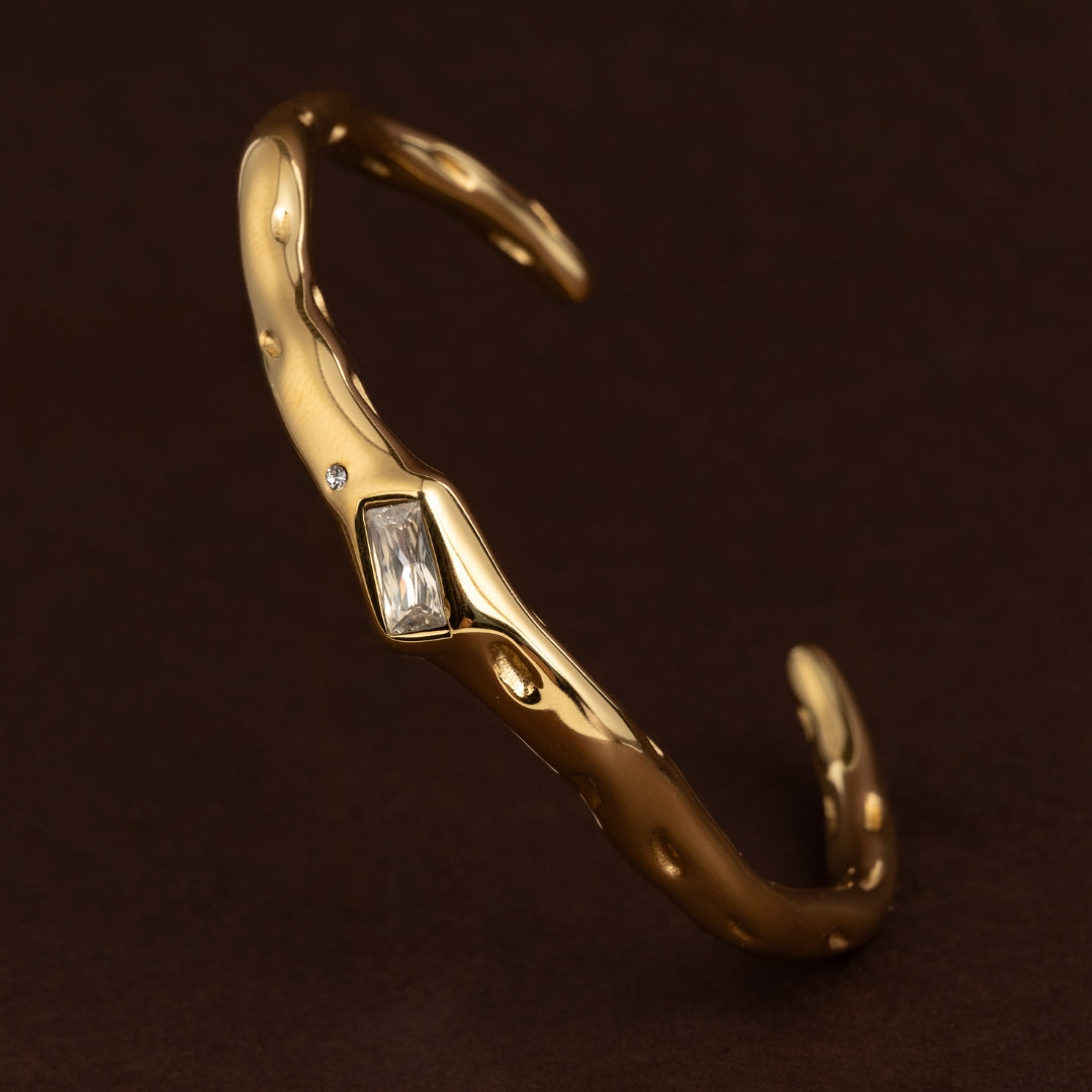 Pulseira escrava dourada aberta (cuff) com textura irregular de metal derretido, cravejada com uma zircónia retangular baguette e um cristal redondo.