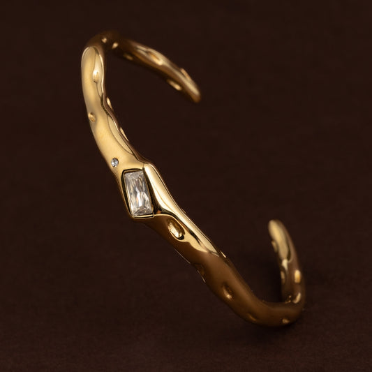 Pulseira escrava dourada aberta (cuff) com textura irregular de metal derretido, cravejada com uma zircónia retangular baguette e um cristal redondo.