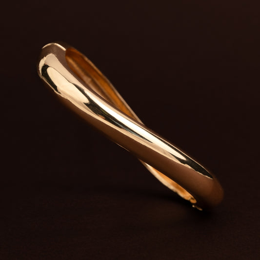 Pulseira rígida dourada com design torcido e fluido, acabamento brilhante sobre fundo escuro