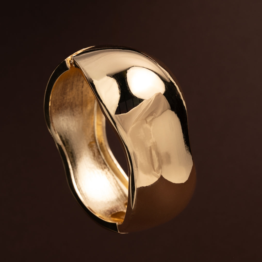 Pulseira escrava larga dourada com acabamento polido e forma orgânica irregular sobre fundo escuro.