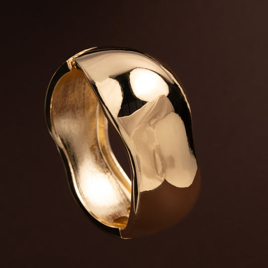 Pulseira escrava larga dourada com acabamento polido e forma orgânica irregular sobre fundo escuro.