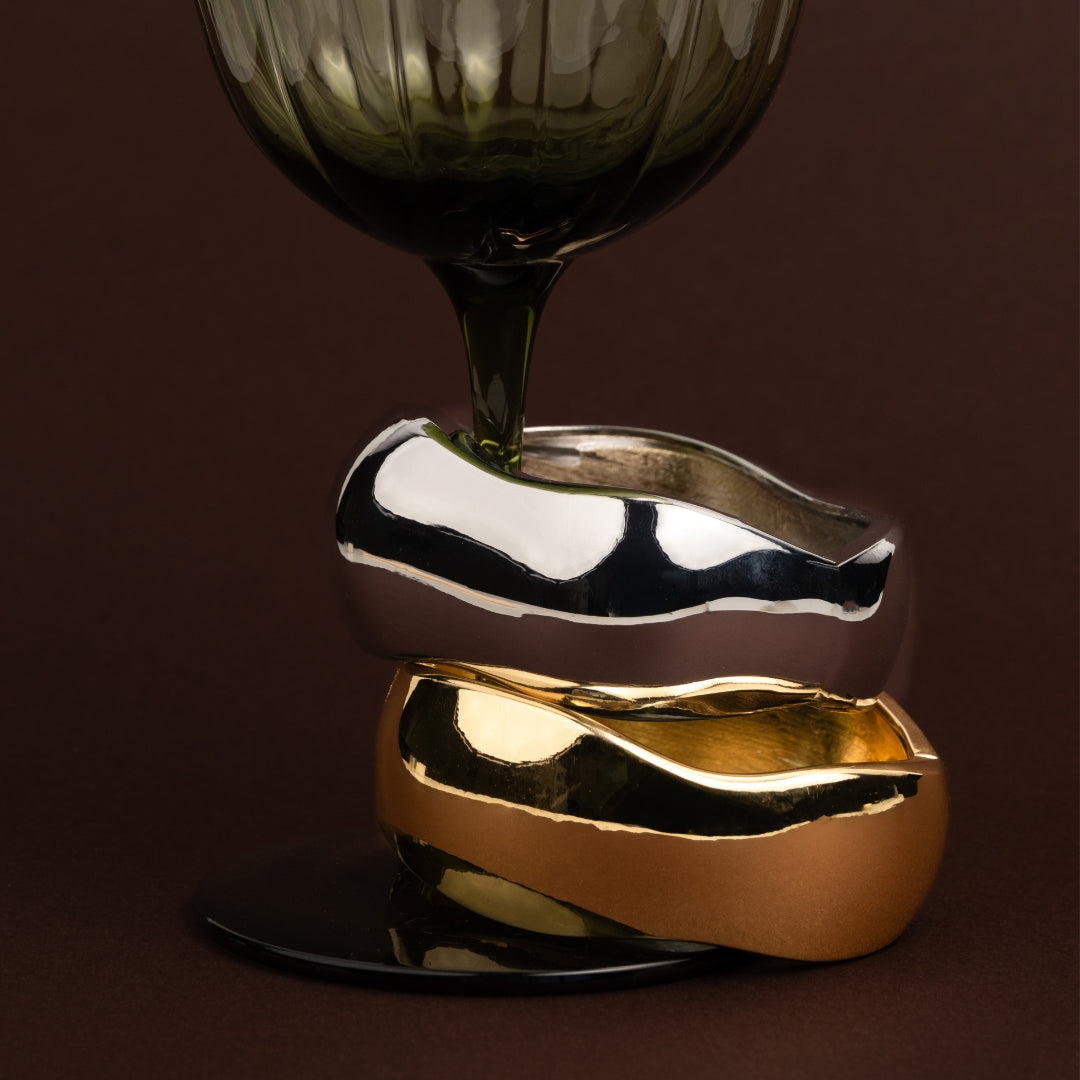 Duas pulseiras largas rígidas estilo escrava, uma dourada e uma prateada, empilhadas, com design ondulado de metal líquido.